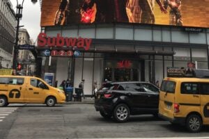 A classic yellow taxi driving past a Seventh Avenue subway entrance in Manhattan, illustrating how to get around NYC.