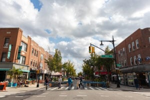 Pedestrians crossing a busy street lined with shops and restaurants in Astoria, Queens, New York.