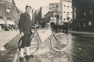 Young messenger boy wearing a uniform and standing beside his bicycle on a wet New York City street in 1896.